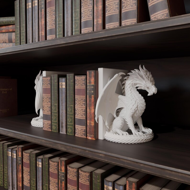 Op de afbeelding: Witte drakenboekensteunen met gedetailleerde vleugels en schubben, geplaatst op een donkere houten plank. De drakenboekensteunen staan tussen een verzameling antieke boeken met verschillende kleuren en texturen. De plank maakt deel uit van een boekenkast.