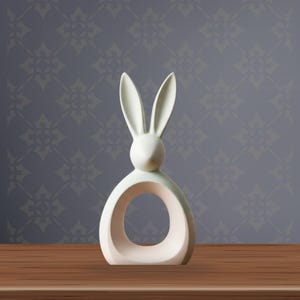 Puede incluir: Una figura de conejo de color crema con orejas largas y una abertura circular en el cuerpo. El conejo está sobre una superficie de madera, con un fondo gris estampado. La figura es un objeto decorativo.