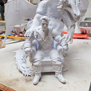 May include: A white 3D printed figurine featuring a person sitting on a throne with a large dragon positioned behind them. The dragon has detailed scales, wings, and sharp claws resting on the shoulders of the person, who wears textured armor.