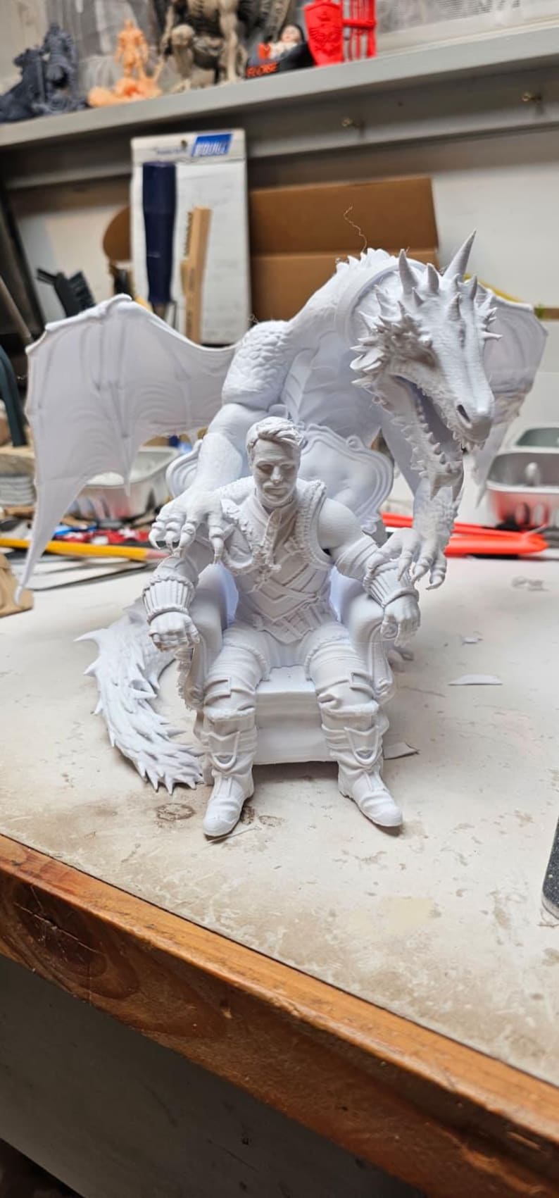 May include: A white 3D printed figurine featuring a person sitting on a throne with a large dragon positioned behind them. The dragon has detailed scales, wings, and sharp claws resting on the shoulders of the person, who wears textured armor.