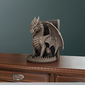 Op de afbeelding: Een bronzen drakenbeeldje met gedetailleerde schubben en vleugels. De draak zit met uitgespreide vleugels. Het is geplaatst naast een donkergrijze boekensteun, waardoor een decoratief fantasie-thema ontstaat.