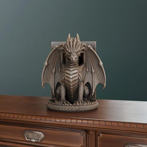 Op de afbeelding: Een gedetailleerd, grijs drakenbeeld met uitgespreide vleugels. De draak zit op een cirkelvormige basis, met zijn vleugels die zijn lichaam omlijsten. Het beeld staat op een houten oppervlak, met een donkergroene achtergrond.