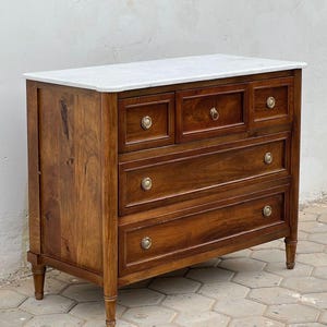 Puede incluir: Una cómoda de madera con una superficie de mármol blanco, tres cajones de ancho completo y dos cajones superiores más pequeños. La pieza está construida en madera de color marrón medio con veta visible y tiradores metálicos redondos.