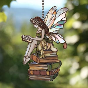 Op de afbeelding: Een gebrandschilderd feeënornament. De fee wordt afgebeeld terwijl ze een boek leest terwijl ze bovenop een stapel boeken zit. De fee heeft bruin haar, een bloemenkrans en iriserende vleugels. Het ornament hangt aan een ketting.