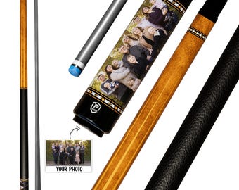 May include: A pool cue featuring a light brown wood grain shaft, a black textured grip, and a custom section displaying a printed family photograph. The cue includes a white and black geometric pattern border and a small logo near the base.
