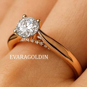 May include: A gold engagement ring with a large, round, clear diamond center stone. The band is smooth and polished, with smaller diamonds set along the sides of the center stone. The ring is on a person's finger.
