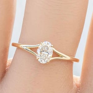 May include: A gold ring with a split shank design, featuring a sparkling oval-cut diamond as the centerpiece. The ring is set against a soft background, highlighting the delicate details of the jewelry. The text "EVARAGOLDIN" is visible.