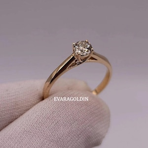 May include: A gold engagement ring with a single, round-cut diamond. The ring has a delicate band and a four-prong setting. The text "EVARAGOLDIN" is visible on the ring.
