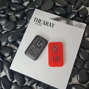 Pode incluir: Dois marcadores retangulares num cartão branco com a etiqueta THE XRAY STUDIO. Um marcador é preto com as letras L e LP, e o outro é vermelho com as letras R e LP. Os marcadores estão colocados num cartão branco sobre pedras pretas.