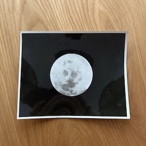 May include: A black and white print depicting the moon. The moon is a detailed circle with craters and texture, set against a black backdrop. The print has a white border and is displayed on a wooden surface.