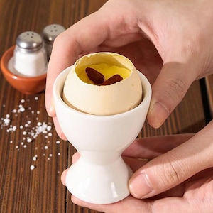 Può includere: Un portauovo in ceramica bianca con un uovo alla coque con tuorlo giallo e guarnizione rosso scuro. Saliere e pepiere sullo sfondo. Il portauovo è tenuto tra le mani.