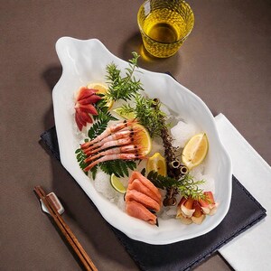 May include: A white, leaf-shaped serving platter filled with sashimi, shrimp, and garnishes. Slices of salmon, lemon wedges, and sprigs of greenery are arranged on a bed of ice. A glass of yellow liquid sits nearby.
