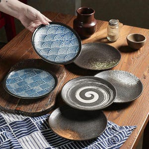 May include: A selection of ceramic plates with diverse patterns. One plate displays a blue and white wave design, another a blue and white grid. Other plates have speckled, spiral, and solid dark finishes. The plates are arranged on a wooden table.