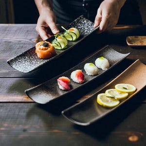 May include: Sushi served on rectangular black ceramic plates. The plates display sushi rolls, including salmon, cucumber rolls, and nigiri. The plates are arranged on a dark wooden surface. A person's hands are holding one of the plates.