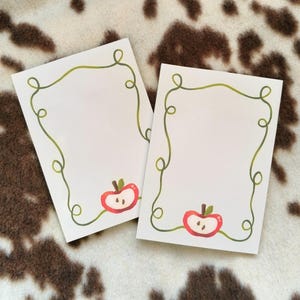 Op de afbeelding: Twee witte rechthoekige kaarten met een groene slingergrens en een rode appelillustratie. De appel is doormidden gesneden en onthult de zaden. De kaarten liggen op een bruin en wit patroon.