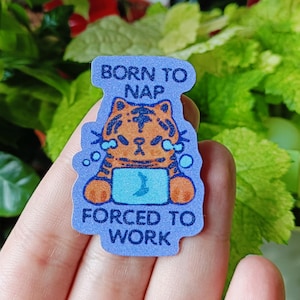 May include: A blue sticker with the text "BORN TO NAP" at the top and "FORCED TO WORK" at the bottom. The sticker features a cartoon tiger with orange and brown stripes, sitting in front of a blue laptop screen. The tiger has a sad expression.