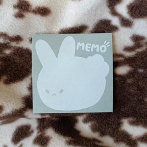 Peut inclure: Un bloc-notes carré vert clair avec un motif de lapin blanc et le mot "MEMO" imprimé en blanc. Le lapin a de grandes oreilles et un petit visage. Le bloc-notes est posé sur une surface à motifs marron et blanc.