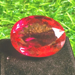 Könnte beinhalten: Ein facettierter, ovaler Edelstein in leuchtendem Rot mit polierter Oberfläche. Der Stein weist zahlreiche geometrische Schliffe auf, die das Licht reflektieren und für ein brillantes Aussehen sorgen. Er liegt auf einem dunklen, strukturierten Sockel.