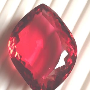 May include: A large, faceted gemstone in a deep red colour. The gem is set against a white, ridged background, highlighting its multiple facets and internal reflections. The overall shape is a modified square with rounded corners.