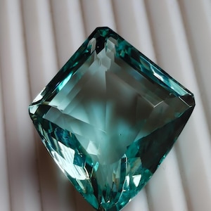 May include: A square-cut, teal-colored gemstone with multiple facets. The gem is a vibrant shade of blue-green, with a polished, reflective surface. The gemstone is set against a white, ridged background, highlighting its clarity and brilliance.