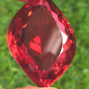 May include: A marquise-cut gemstone with a vibrant, translucent red color and numerous geometric facets that reflect light. The stone features a pointed shape at both the top and bottom ends, showcasing a clear, polished surface throughout.