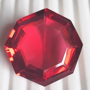 以下が含まれることがあります： 六角形にカットされた、透明な赤いファセットカットの宝石。石には光を反射する複数の平らで磨かれた表面があり、幾何学的な構造全体に深いクリムゾンレッドから明るい赤までの様々な色合いを作り出しています。