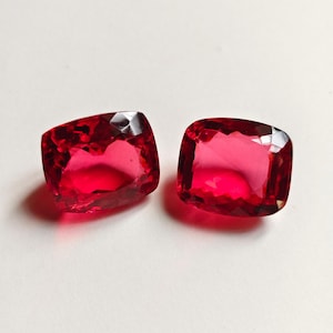 May include: Two loose cushion-cut gemstones with a vibrant, deep red color. Each stone features a faceted surface that reflects light, creating a brilliant appearance. The gems are positioned side by side, showcasing their symmetrical shape.