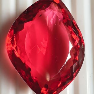 Peut inclure: Une pierre précieuse rouge translucide et vibrante, dotée d'une forme rappelant la marquise et de facettes complexes. La pierre présente des teintes cramoisies profondes et des bords géométriques nets qui reflètent la lumière.