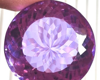 May include: A round, faceted purple gemstone featuring a complex, symmetrical geometric pattern cut into its center. The stone displays varying shades of violet and lavender, with sharp, reflective edges throughout its circular surface.