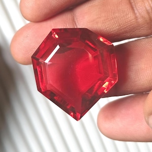 May include: A faceted, octagonal, deep red gemstone. The gem is held in a hand, showcasing its clarity and vibrant color. The cut of the stone creates multiple reflective surfaces, enhancing its brilliance. The gemstone appears to be a ruby or similar precious stone.