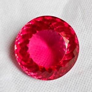 May include: A large, round, faceted gemstone in a vivid red hue. The gem displays a high degree of clarity and brilliance, with numerous facets reflecting light. The gemstone is set against a white background.