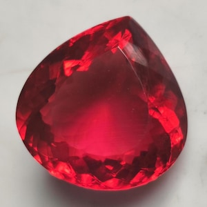 May include: A faceted, teardrop-shaped gemstone in a vibrant shade of red. The gem's surface reflects light, showcasing its intricate cuts and internal structure. The gemstone is a striking example of a precious stone.