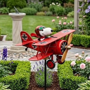 May include: A red metal airplane yard decoration featuring a cartoon dog character wearing a pilot's helmet and goggles. The biplane has a wooden propeller and black crosses on the wings. The garden ornament is mounted on a black metal stake.