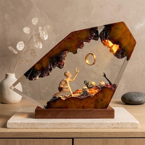 Könnte beinhalten: Harzskulptur mit einem goldenen Ring, der über einer Szene mit einer knienden Figur, Schädeln und einem feurigen Hintergrund schwebt. Das Kunstwerk ist in einer transparenten, geometrischen Form eingeschlossen und ruht auf einem Holzsockel.