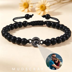 Puede incluir: Una pulsera de cuentas negras con esferas mate y un dije circular central. El dije contiene una lente de proyección que muestra una fotografía circular de dos personas abrazándose al aire libre. La pulsera tiene un cierre de cordón ajustable.