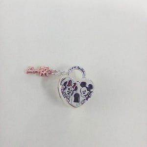 May include: A silver heart-shaped padlock charm with a rose gold key. The padlock features engraved images of Minnie and Mickey Mouse. The key is attached to the padlock with a small chain. The charm is on a white background.