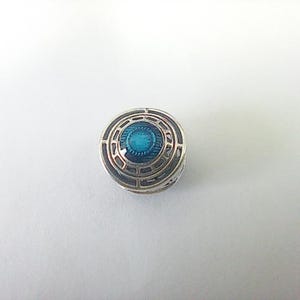 Può includere: Un ciondolo circolare color argento con un design geometrico dettagliato. Il centro presenta un'incastonatura in smalto di colore turchese vibrante. Il ciondolo ha un aspetto a strati, con una serie di cerchi concentrici e una superficie strutturata.