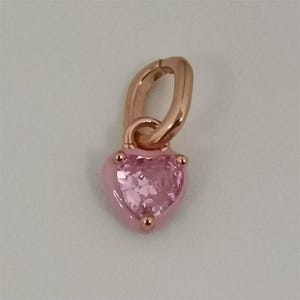 May include: A rose gold charm with a pink heart-shaped gemstone. The charm has a hinged clasp for easy attachment. The heart is surrounded by a pink enamel border and has three small decorative studs.