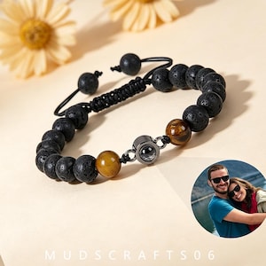 Puede incluir: Una pulsera de cuentas con piedras de lava negra porosas y dos cuentas lisas de ojo de tigre marrón dorado. El centro incluye un pequeño dije metálico circular de proyección. La pulsera tiene un cordón negro con un cierre de nudo corredizo.