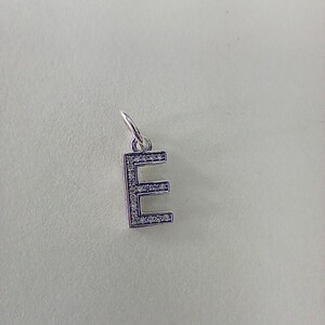 May include: A silver-colored charm in the shape of the letter "E". The charm is encrusted with small, clear gemstones along the edges. It has a small loop at the top for attaching to a bracelet or necklace.