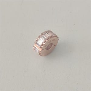 Op de afbeelding: Roségoudkleurige bedel met een rij rechthoekige, heldere stenen. De bedel is een clip-on model, met de tekst "ALE MET" zichtbaar. De bedel is ontworpen voor gebruik met een armband of ketting.