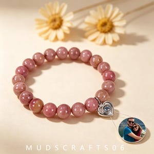 Puede incluir: Una pulsera de cuentas hecha de piedras lisas y redondas de color rosa polvoriento. Un dije plateado en forma de corazón está unido a la pulsera y proyecta una fotografía circular de una pareja abrazándose al aire libre cerca del agua.