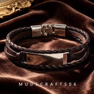 May include: A multi-strand bracelet featuring dark brown braided leather cords and a smooth, polished silver-tone metal ID plate at the center. The bracelet includes a secure silver-tone metal clasp closure. The text MUDSCRAFTS06 is visible.