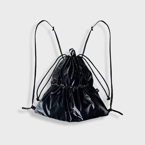 May include: Black drawstring backpack with a glossy, reflective finish. The bag features black drawstrings that function as shoulder straps. The top of the bag is gathered, and it has a small tag on the side. Designed to be worn on the back.