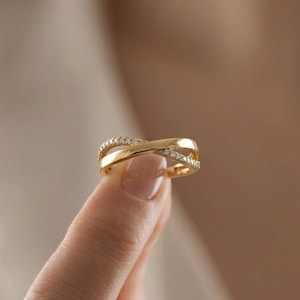 May include: A gold ring with a crisscross design, embellished with small, sparkling stones. The ring is held up by a finger, highlighting its intricate details and elegant style. The ring's design features a band that crosses over itself, creating a unique and eye-catching look.