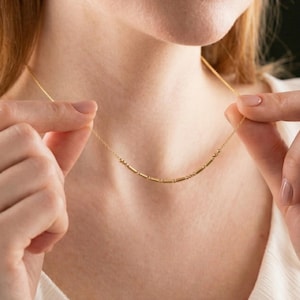 Puede incluir: Un delicado collar de oro con una serie de pequeñas cuentas espaciadas uniformemente. El collar lo sostiene una persona que lleva una blusa blanca. El collar es una joya sencilla y elegante.