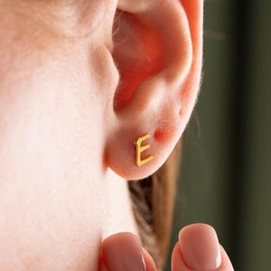 Może przedstawiać: Zbliżenie ucha z małą, złotą kolczykiem w kształcie litery "E". Kolczyk znajduje się na płatku ucha. Karnacja jest jasna, a paznokcie pomalowane są jasnoróżowym lakierem. Tło jest rozmyte zielone.