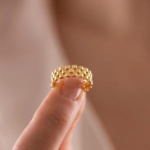 May include: A gold ring featuring a rectangular link pattern. The ring is held between two fingers. The design consists of a repeating pattern of small, rectangular links, creating a textured surface. The ring has a bright, shiny gold colour.