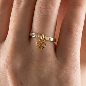 May include: A gold ring with a heart-shaped charm. The ring band is adorned with small, clear stones. The heart charm hangs from a small bar attached to the ring. The ring is on a finger.