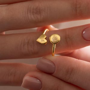 Könnte beinhalten: Ein goldfarbener Ring mit einem Herz- und einem kreisförmigen Design. Der Ring ist offen, so dass er an verschiedene Fingergrößen angepasst werden kann. Der Ring wird an einem Finger gezeigt.
